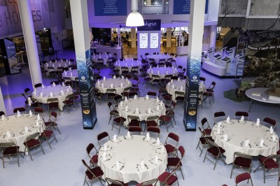 Salle lumineuse avec des tables en bois, des stands d'exposition et des personnes observant ou manipulant des objets.