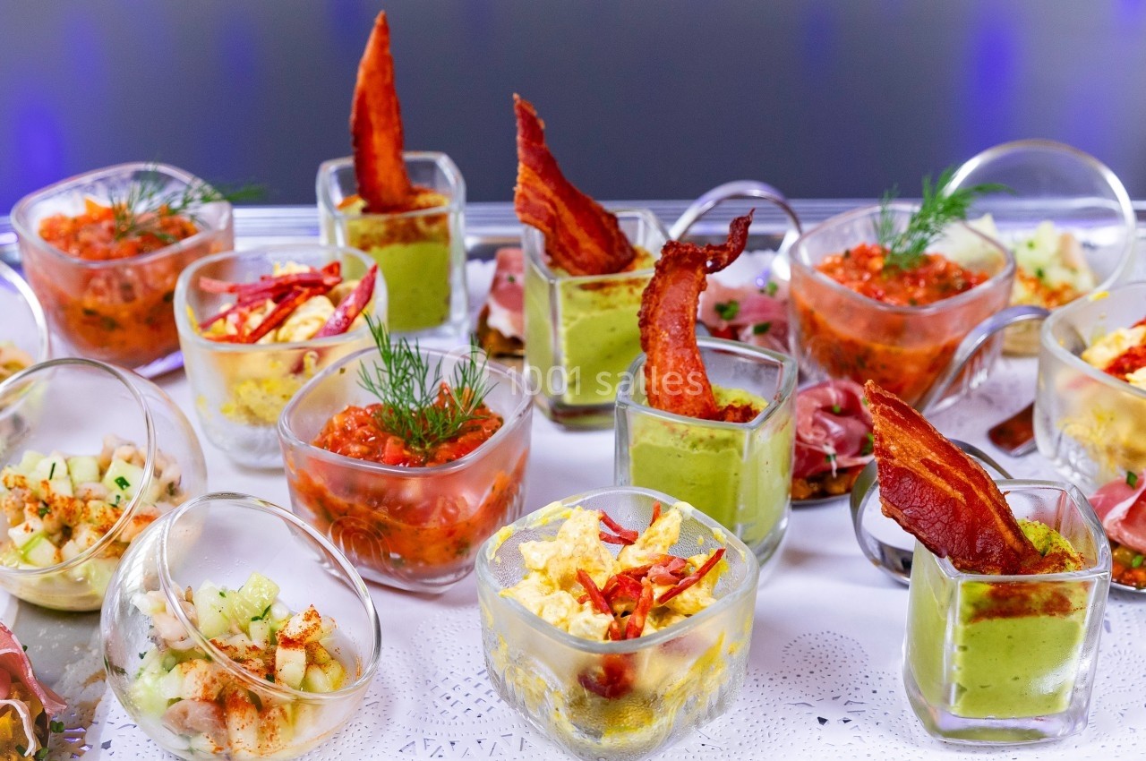 Assortiment de verrines colorées garnies de légumes, sauces, bacon et herbes fraîches, présentées sur une nappe blanche.