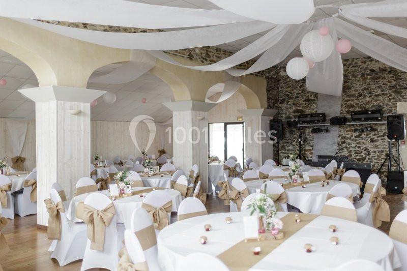 Salle de réception décorée avec des tables rondes, nappes blanches, chaises ornées de nœuds beige et guirlandes suspendues.