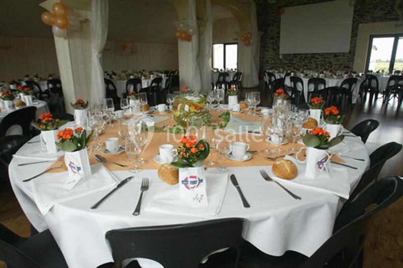 Salle de réception décorée avec des tables rondes dressées, nappes blanches, fleurs orange et vaisselle élégante.