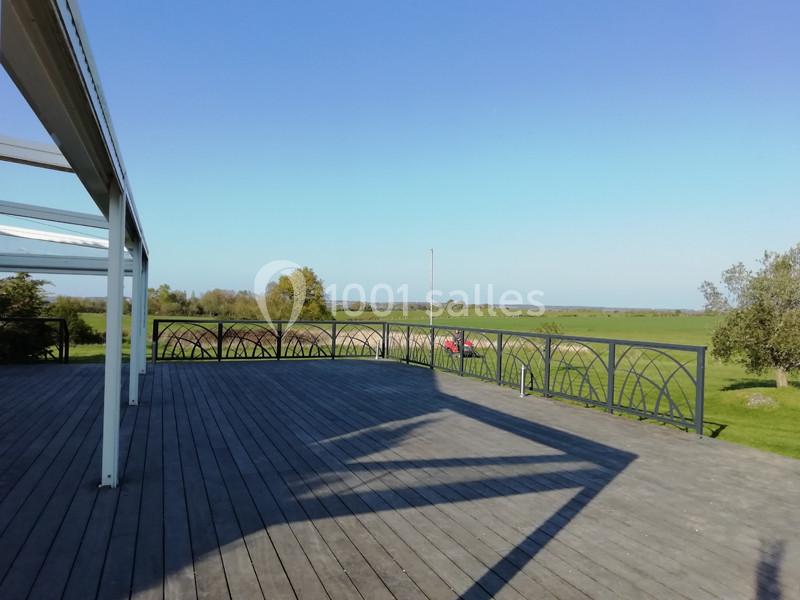 Terrasse en bois avec garde-corps métallique, offrant une vue dégagée sur un paysage champêtre sous un ciel bleu.