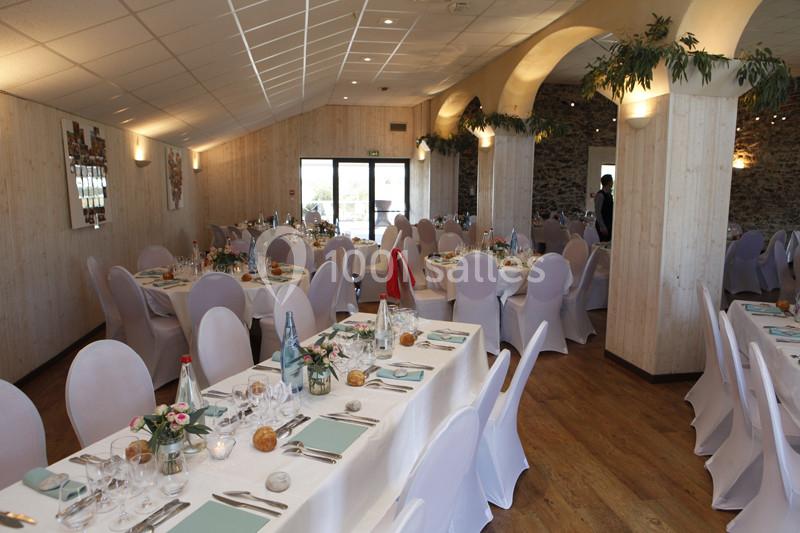 Salle de réception décorée avec des tables dressées, nappes blanches, chaises houssées et lumière naturelle.