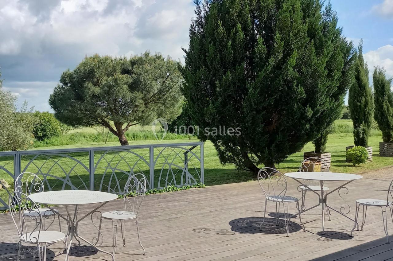 Terrasse en bois avec tables et chaises en métal, entourée d'arbres et donnant sur un espace vert.