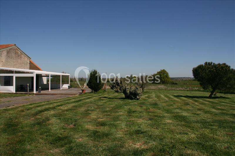 Jardin avec pelouse bien entretenue, arbres dispersés et une maison avec terrasse couverte à gauche sous un ciel dégagé.