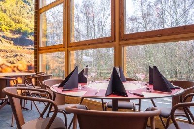Salle de restaurant lumineuse avec de grandes baies vitrées, des tables en bois et des chaises marron.