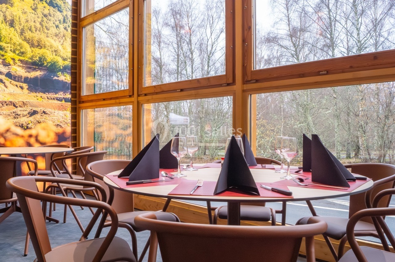 Salle de restaurant lumineuse avec grandes baies vitrées, tables dressées et vue sur un paysage boisé.