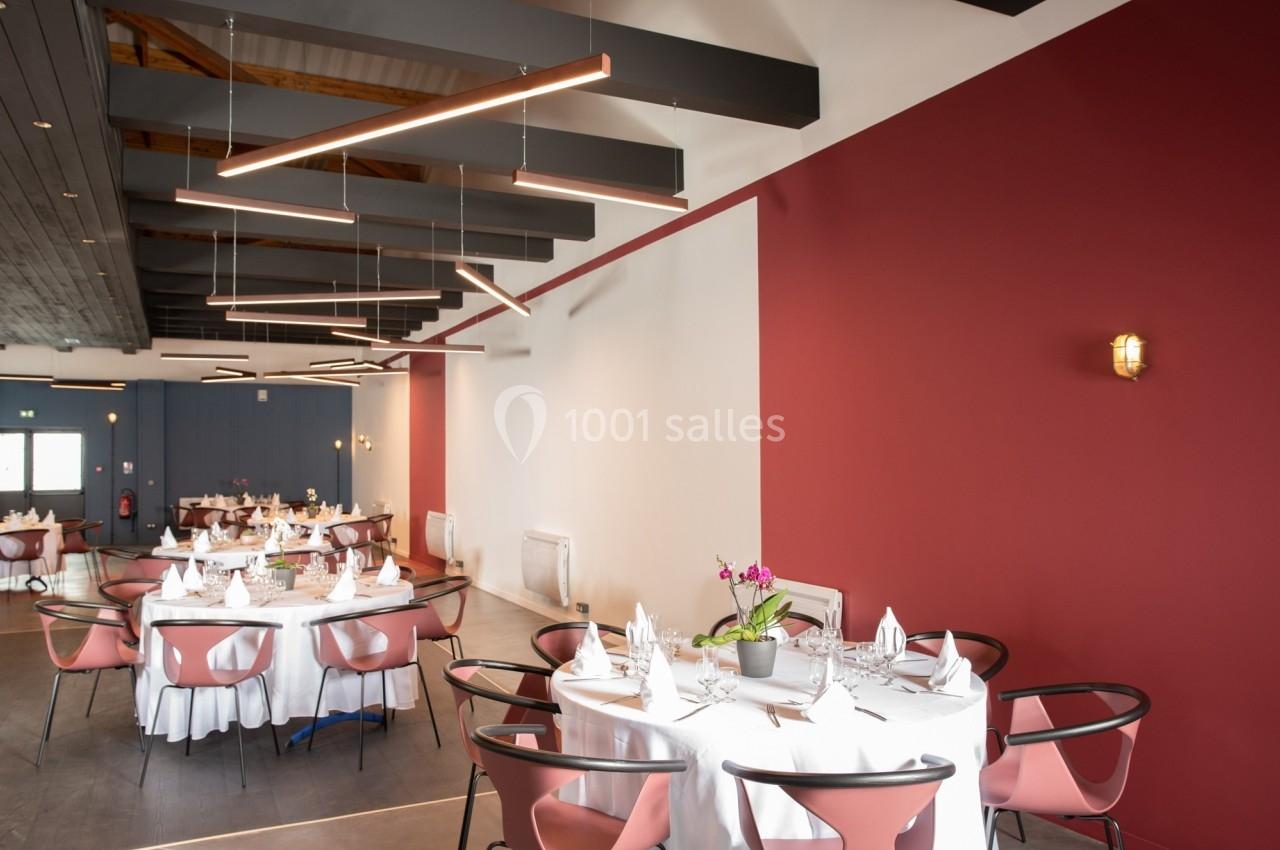 Salle de réception moderne avec tables rondes dressées, chaises roses et murs rouges et blancs.