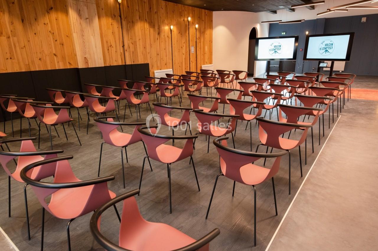 Salle de conférence moderne avec des rangées de chaises rouges et deux écrans affichant un logo à l'arrière-plan.