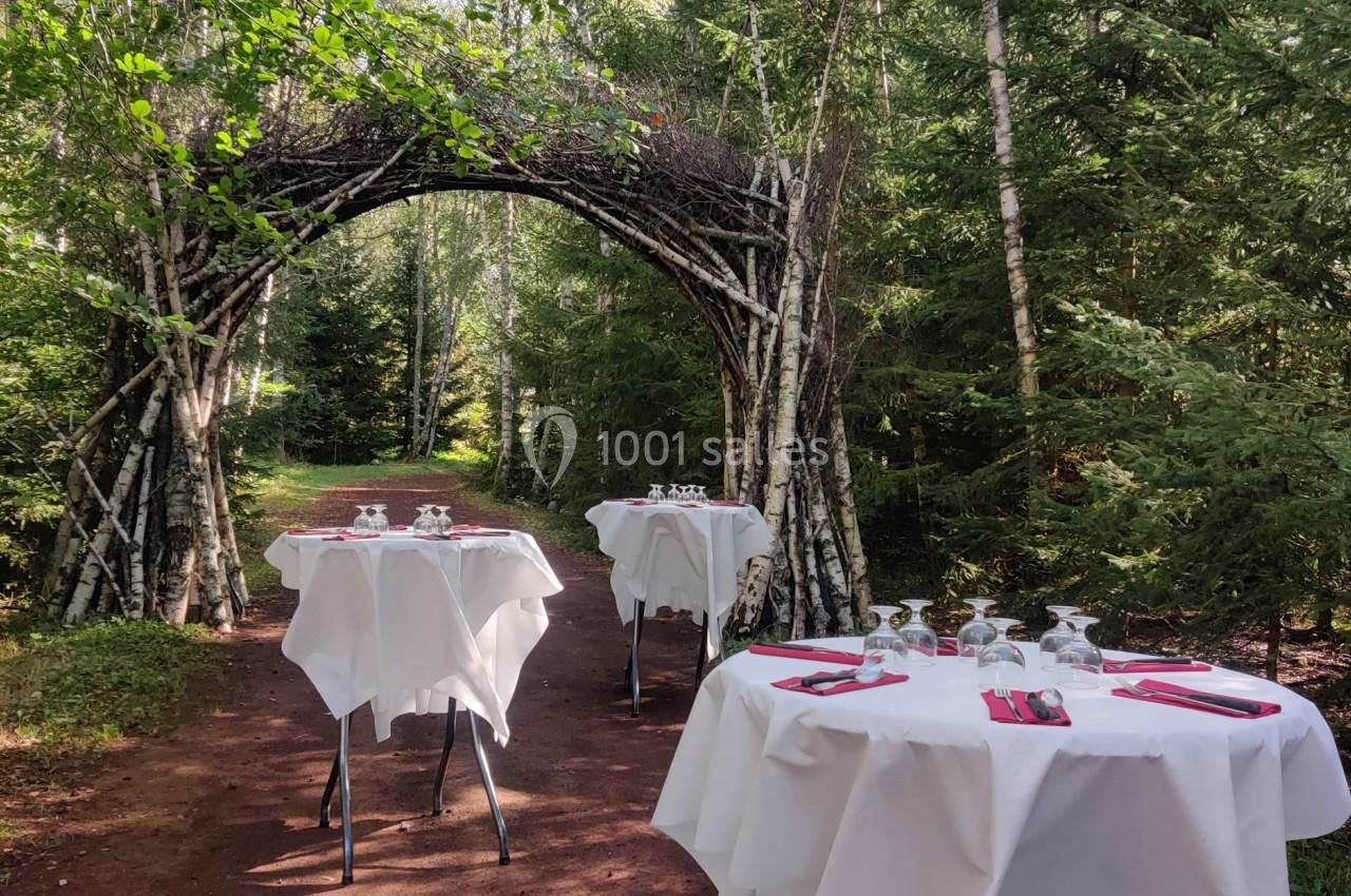 Tables dressées avec nappes blanches et couverts, disposées en extérieur sur un chemin forestier sous une arche en bois.