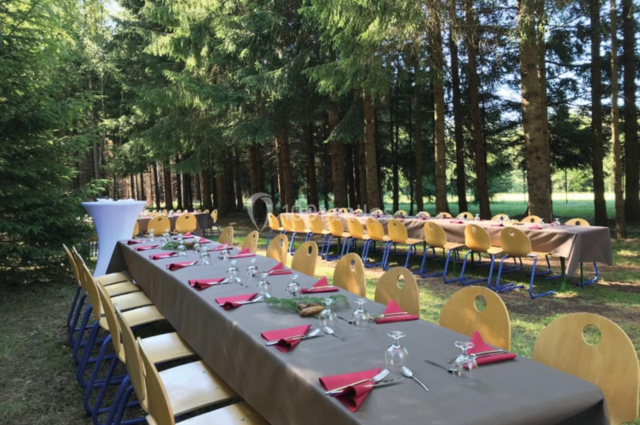 Tables dressées avec nappes brunes, serviettes rouges et verres, disposées en extérieur dans une forêt.