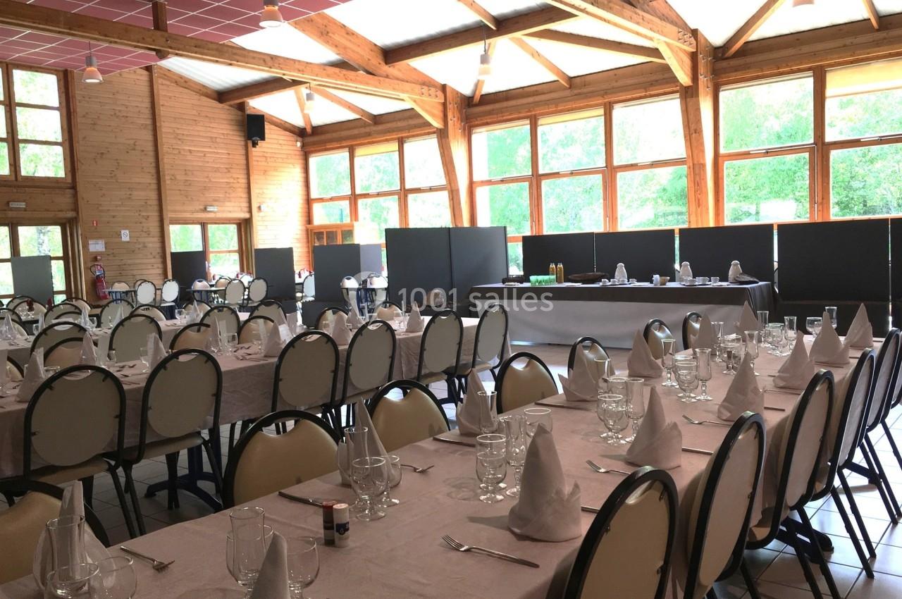 Salle lumineuse avec tables dressées pour un repas, nappes blanches, serviettes pliées et grandes baies vitrées.