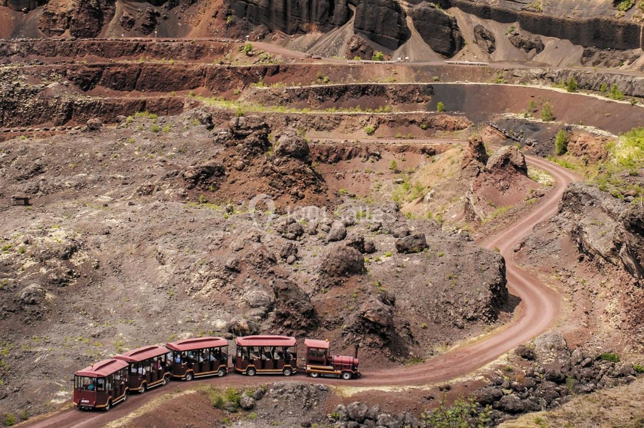 Petit train touristique rouge transportant des passagers dans un paysage volcanique rocheux et aride.