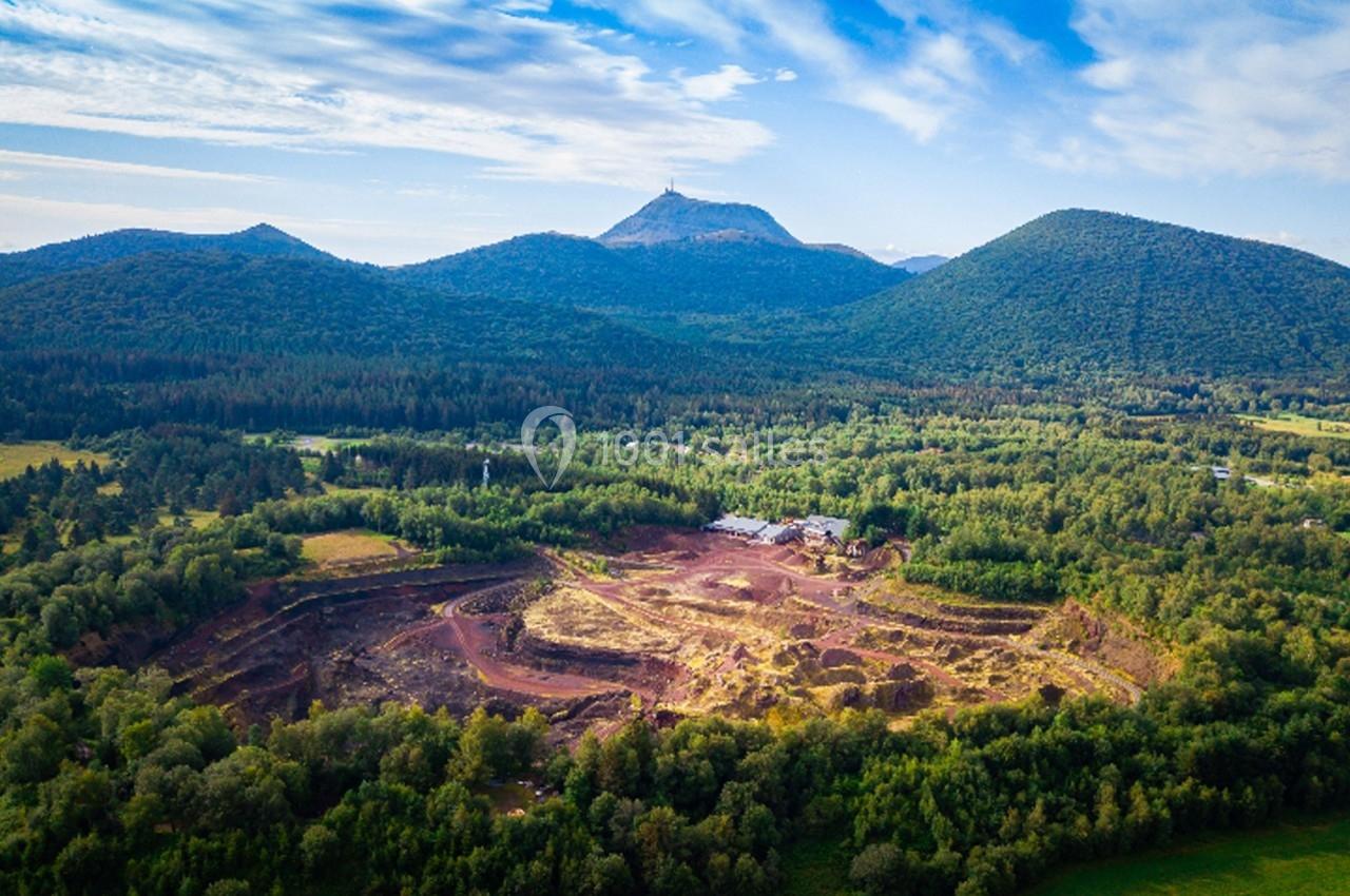Vue aérienne d'une carrière entourée de forêts avec des montagnes en arrière-plan sous un ciel partiellement nuageux.