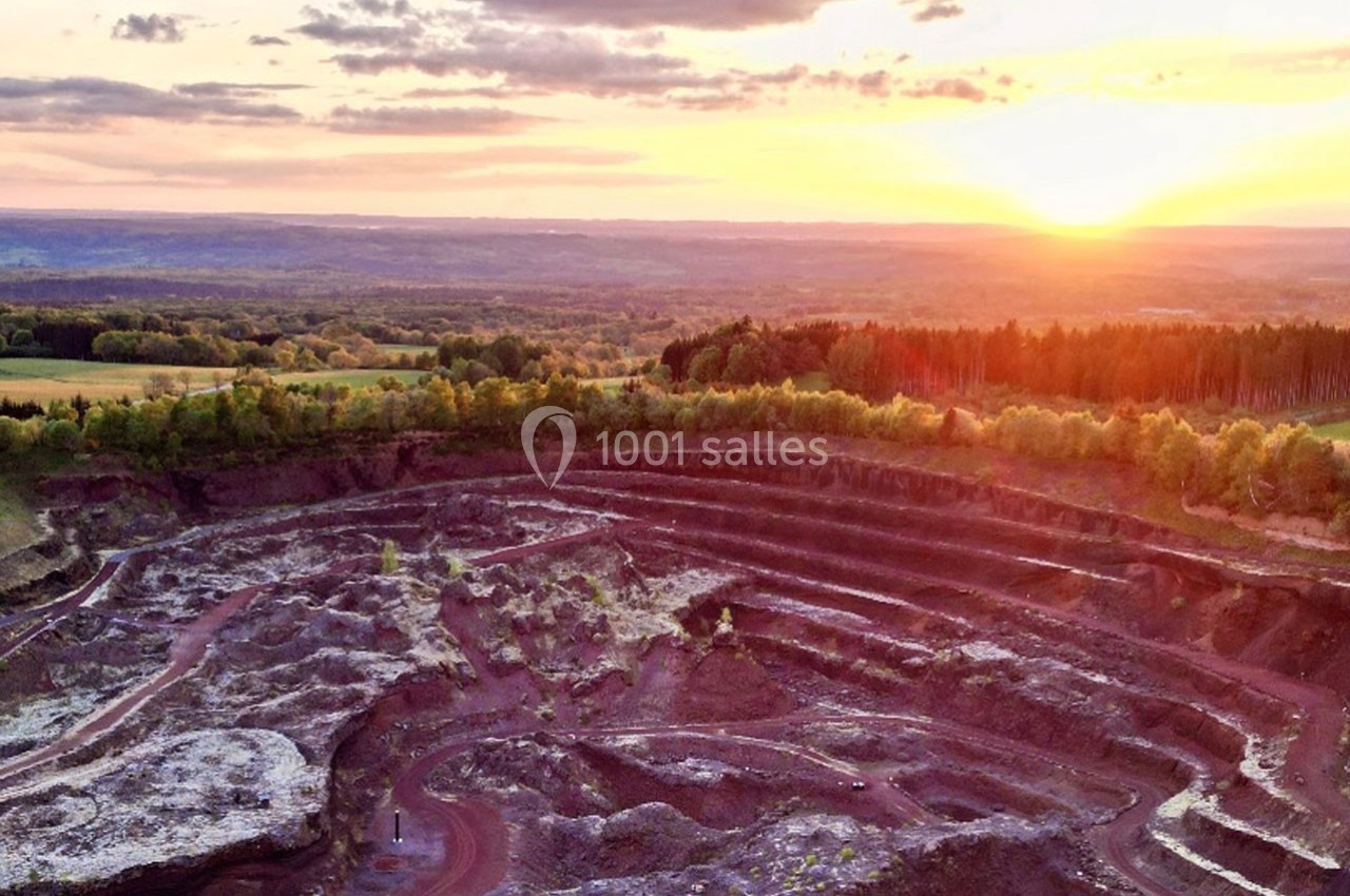 Vue aérienne d'une carrière au coucher du soleil, entourée de forêts et de champs sous un ciel partiellement nuageux.