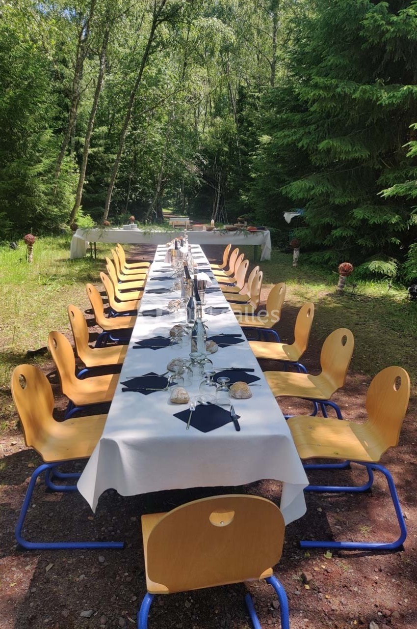Table dressée avec nappes blanches, verres et serviettes noires, entourée de chaises en bois, dans une clairière boisée.