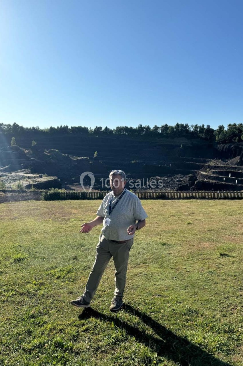 Un homme debout sur une pelouse ensoleillée, avec une carrière et des arbres en arrière-plan.