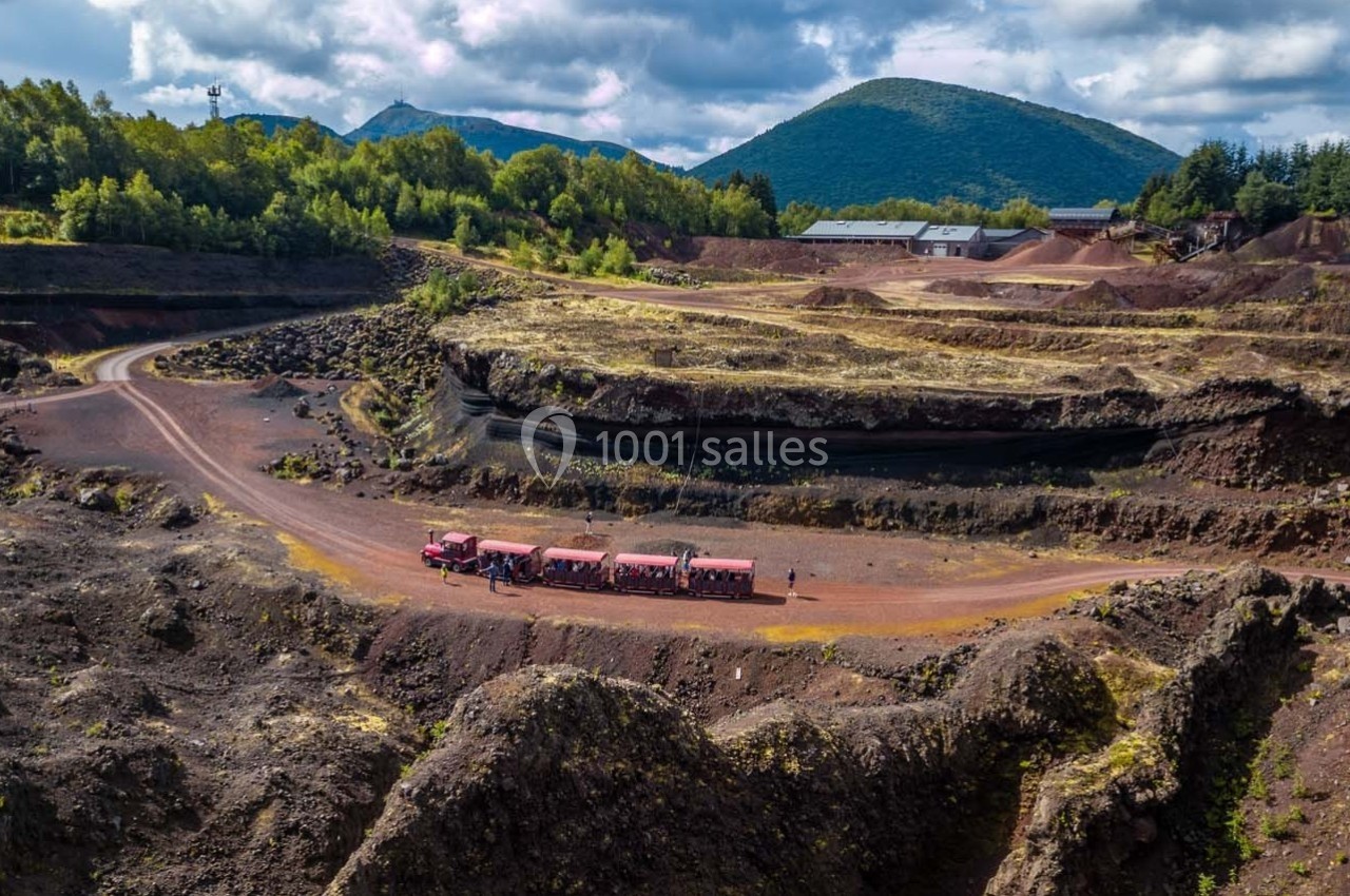 Vue d'une ancienne carrière volcanique avec un petit train touristique rouge et des collines verdoyantes en arrière-plan.