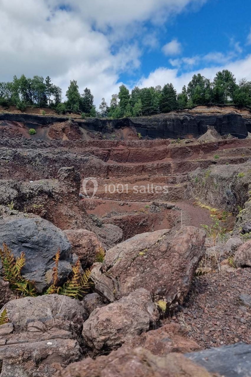 Paysage de carrière avec des roches rouges en terrasses, entouré de végétation et surmonté d'arbres sous un ciel bleu.