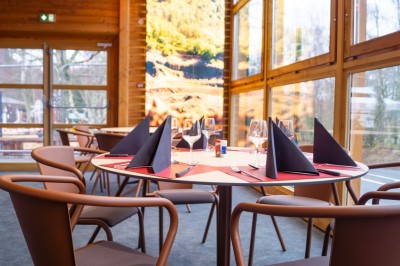 Salle de restaurant lumineuse avec de grandes baies vitrées, des tables en bois et des chaises marron.