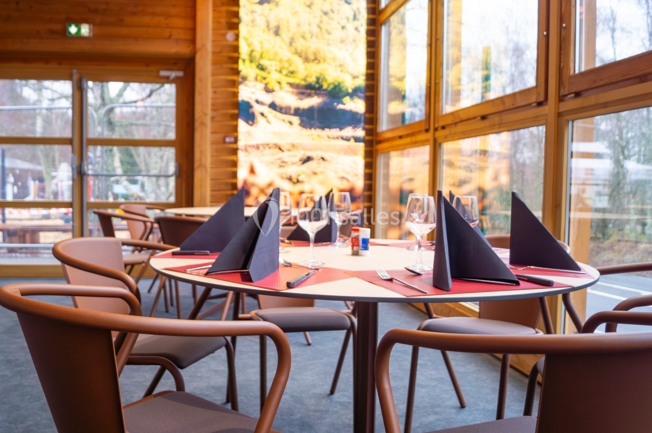 Salle de restaurant lumineuse avec une table dressée, nappes rouges, serviettes noires et grandes baies vitrées.