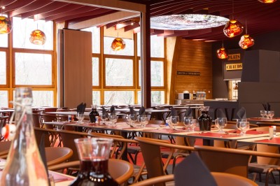 Salle de restaurant lumineuse avec de grandes baies vitrées, des tables en bois et des chaises marron.