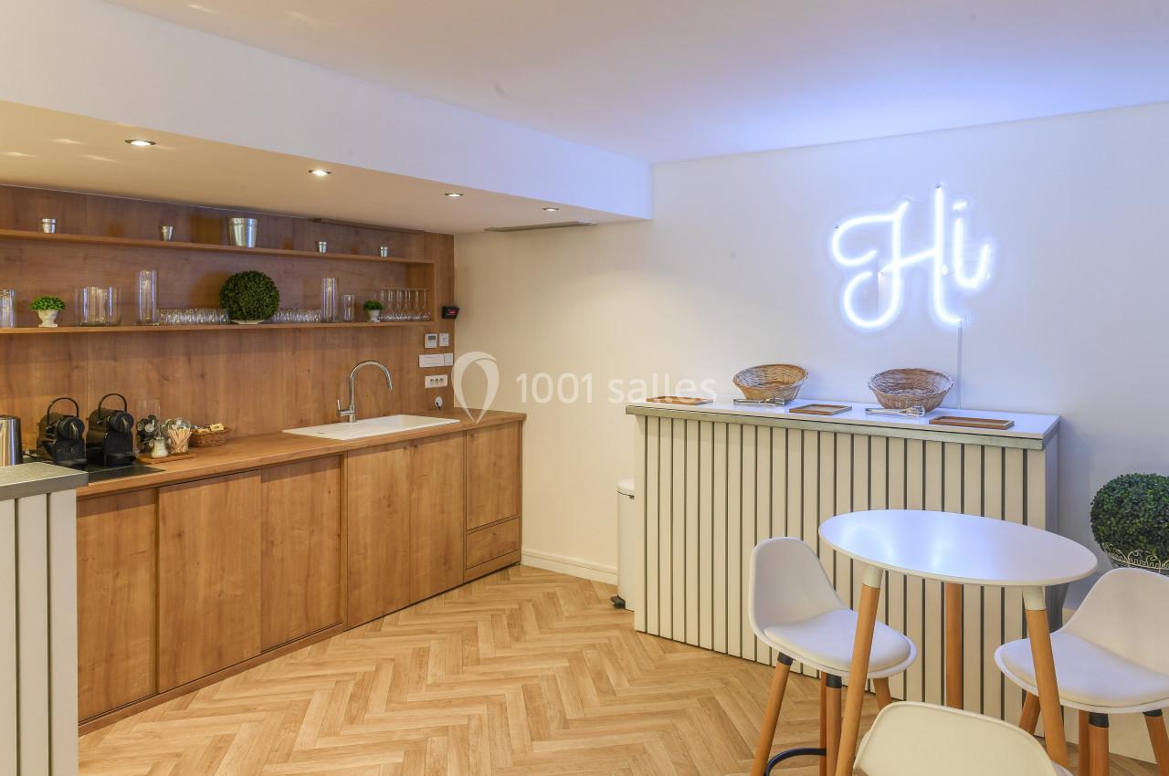 Cuisine moderne avec meubles en bois, comptoir blanc, table ronde et enseigne lumineuse ’Hi’ sur un mur blanc.