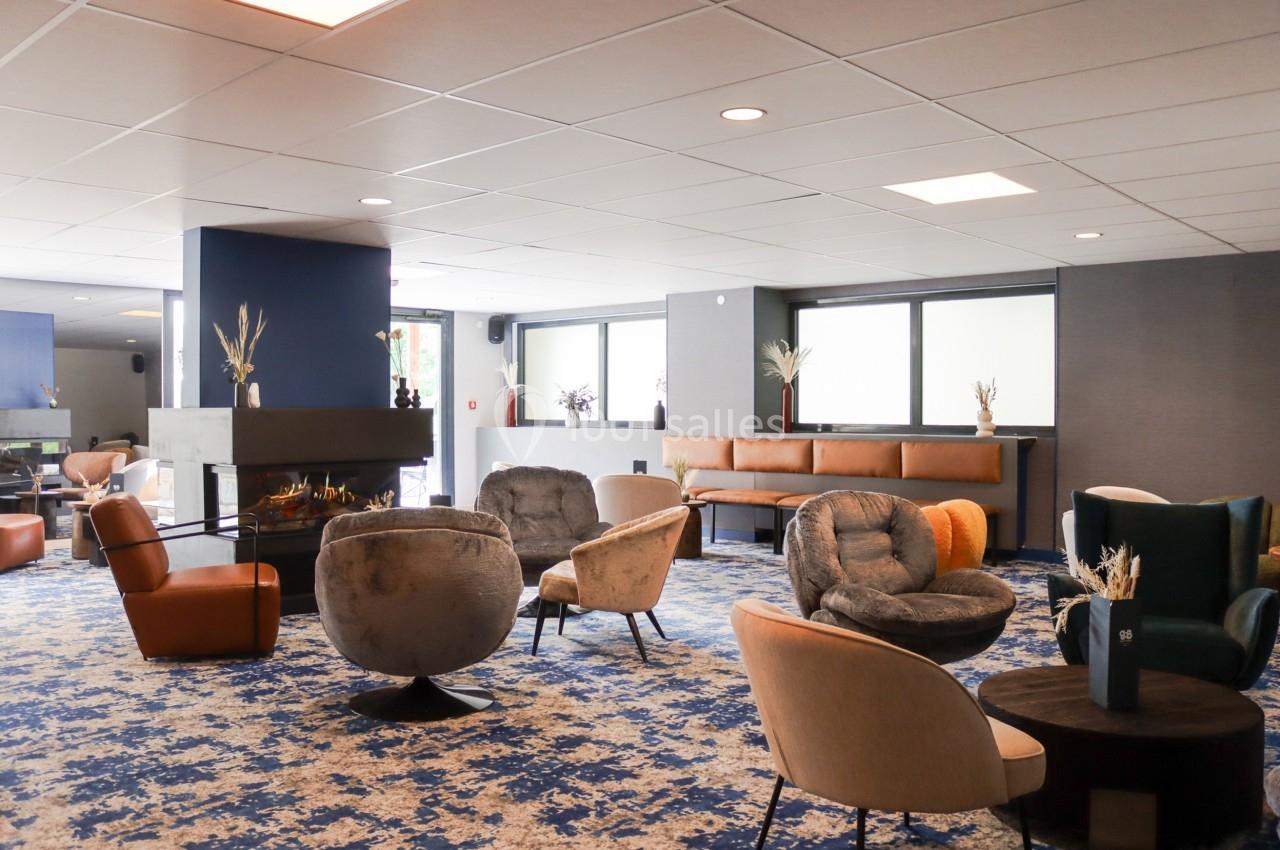 Espace salon moderne avec fauteuils variés, cheminée centrale et grandes fenêtres laissant entrer la lumière naturelle.