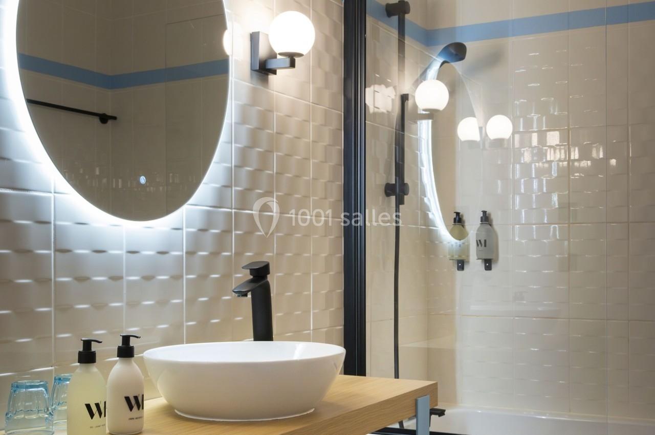 Salle de bain moderne avec lavabo blanc, miroir rond éclairé, robinet noir et carrelage mural texturé.