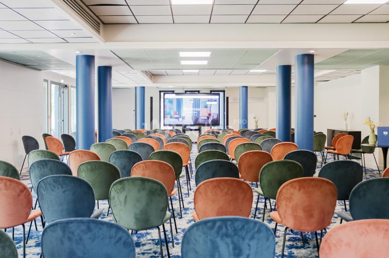 Salle de conférence lumineuse avec des rangées de chaises colorées face à un écran de présentation.