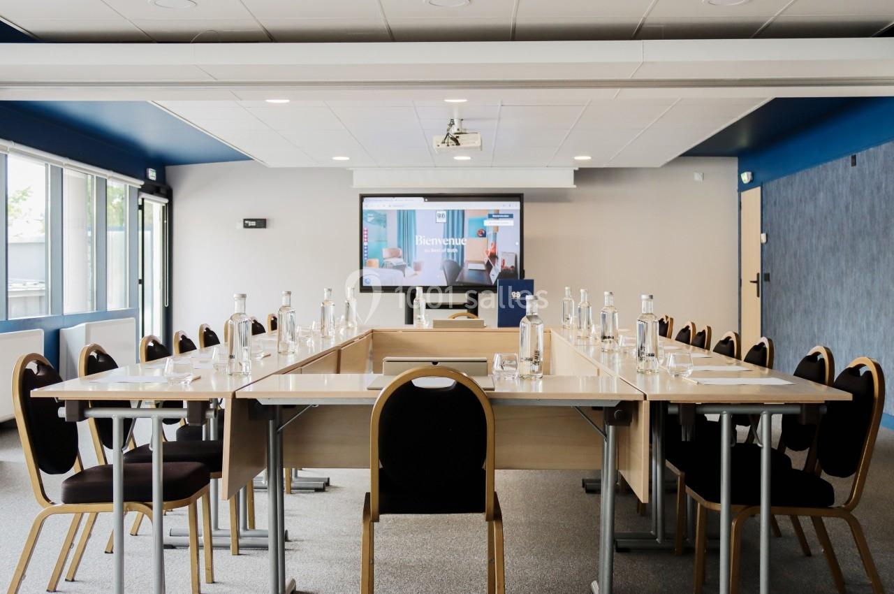 Salle de réunion lumineuse avec tables disposées en U, chaises noires et écran de projection allumé en fond.