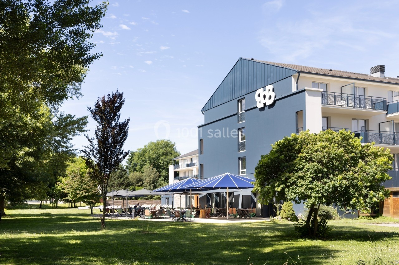 Bâtiment bleu avec terrasses et parasols, entouré d'arbres et d'une pelouse sous un ciel dégagé.