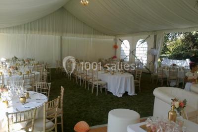 Intérieur d'une tente de réception avec tables rondes décorées, chaises en bois clair et lumière naturelle.