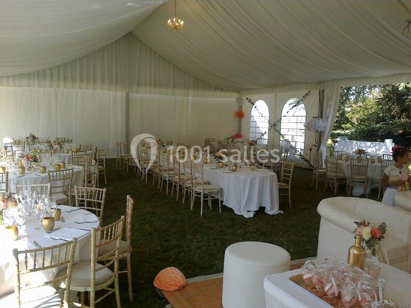 Intérieur d'une tente de réception avec tables rondes décorées, chaises en bois clair et lumière naturelle.