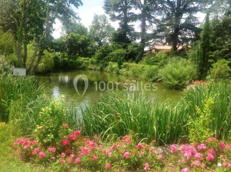 Étang entouré de roseaux, fleurs roses et arbres, avec des canards nageant sur l'eau calme.