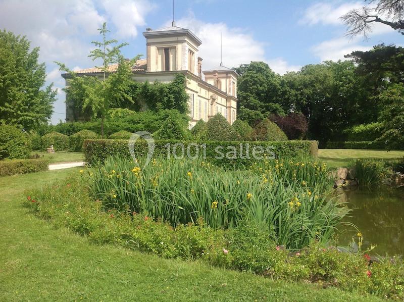 Grande maison entourée de jardins verdoyants, avec un étang bordé de plantes et des arbres en arrière-plan.