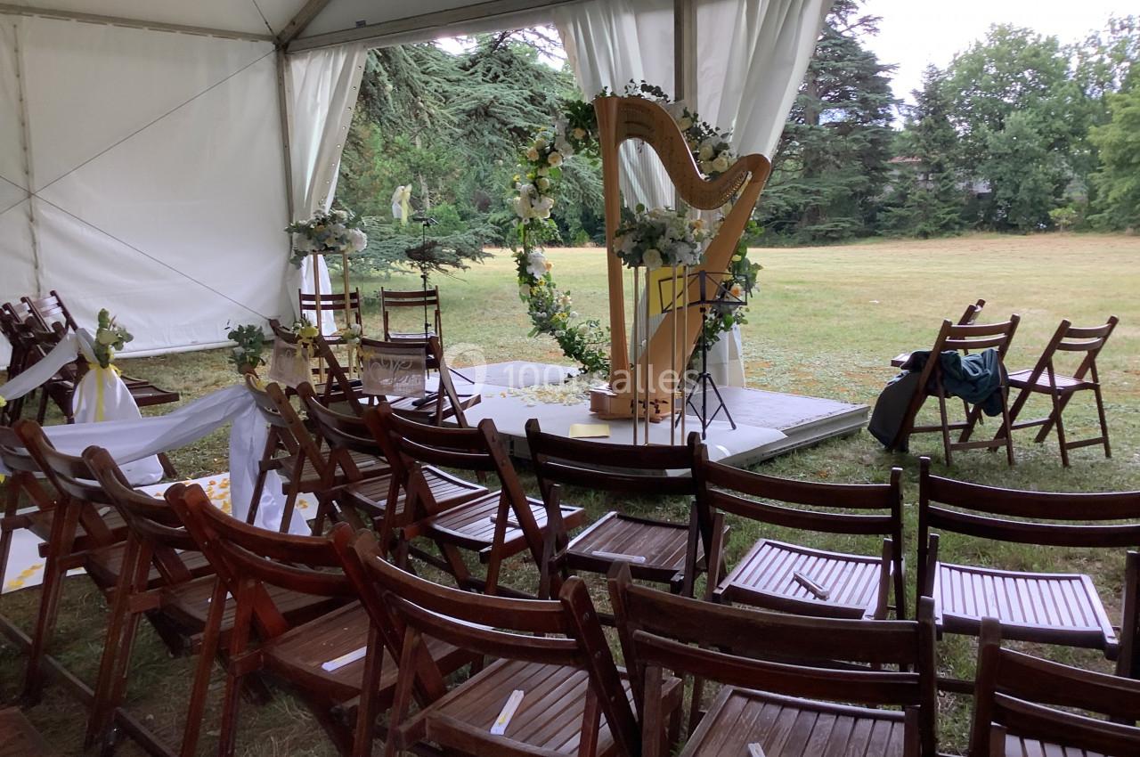 Chaises en bois disposées sous une tente face à une harpe décorée de fleurs, dans un cadre extérieur verdoyant.
