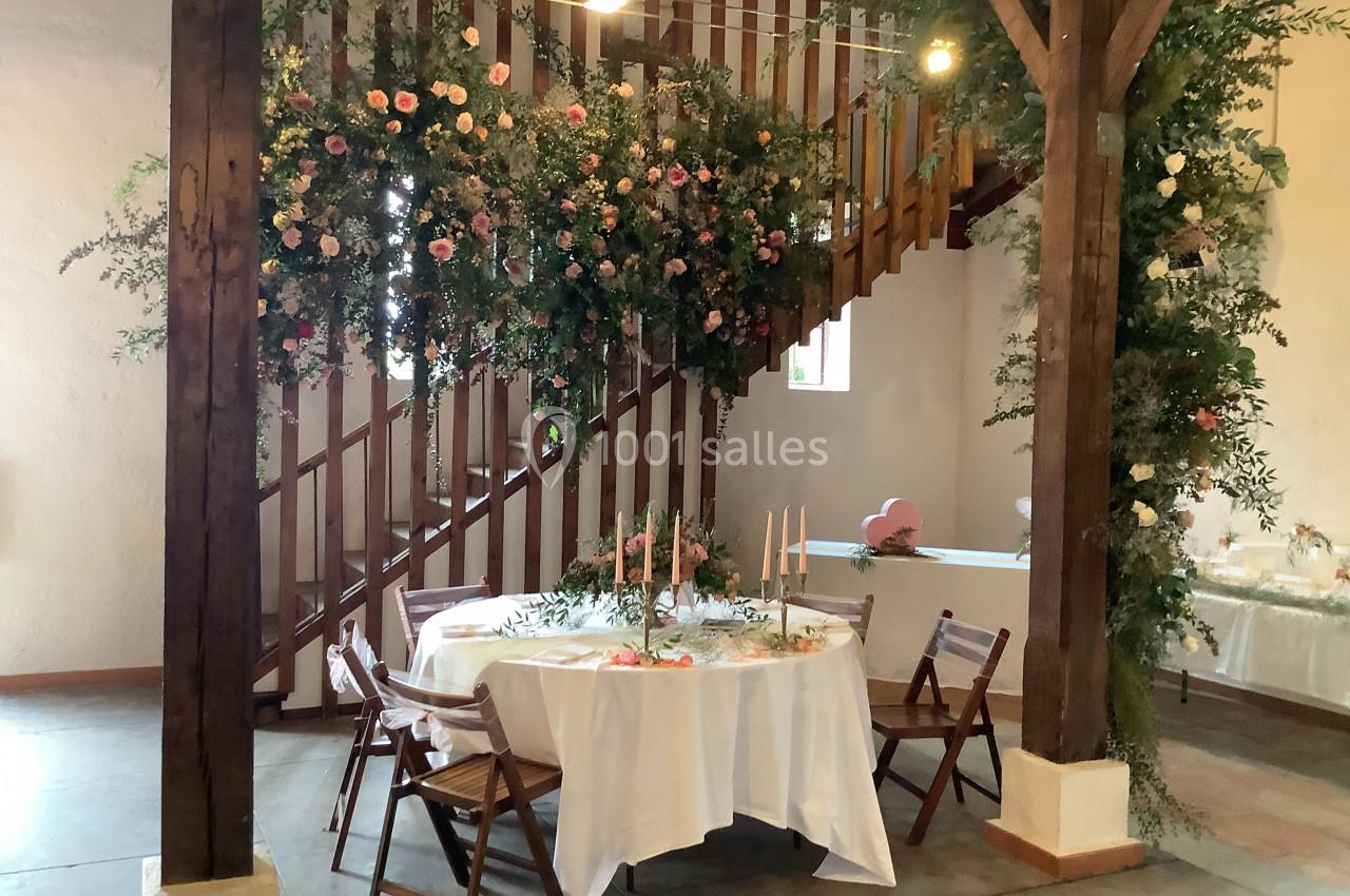 Table ronde décorée de bougies et fleurs sous un escalier en bois, avec une suspension florale et chaises en bois.