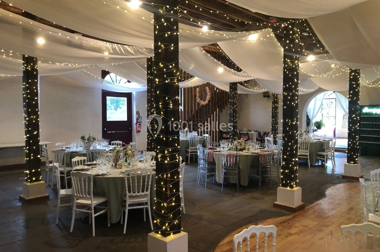 Salle de réception décorée avec des guirlandes lumineuses, des tables rondes dressées et un plafond drapé de voilages blancs.