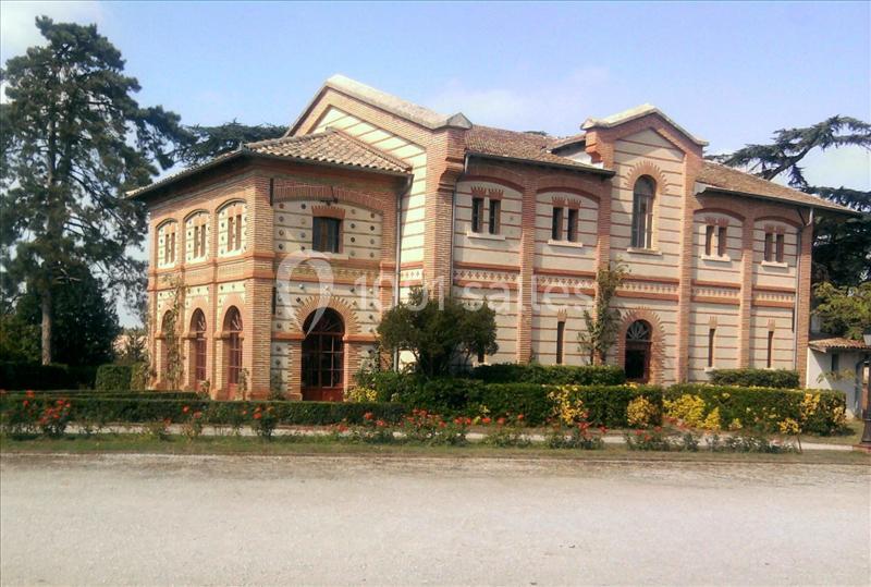 Maison en briques rouges et blanches avec des arches, entourée de jardins et d'arbres, sous un ciel dégagé.