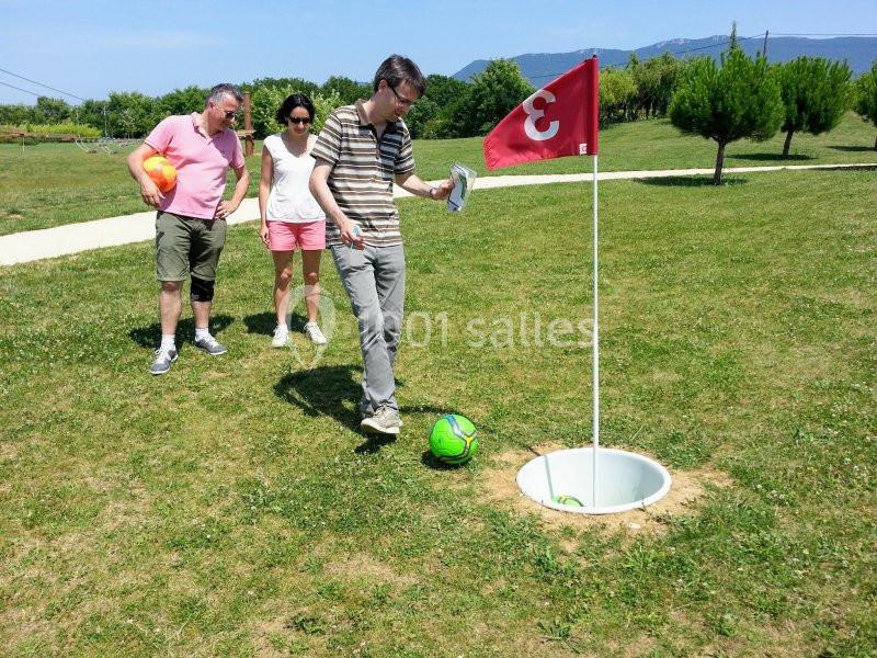 Un homme joue au footgolf en visant un trou, observé par deux personnes sur un terrain herbeux.