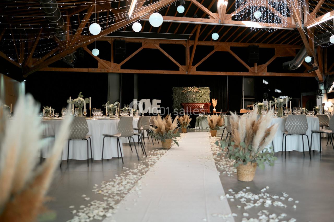 Salle de réception décorée pour un mariage, avec tables dressées, allée centrale ornée de pétales et éléments végétaux.