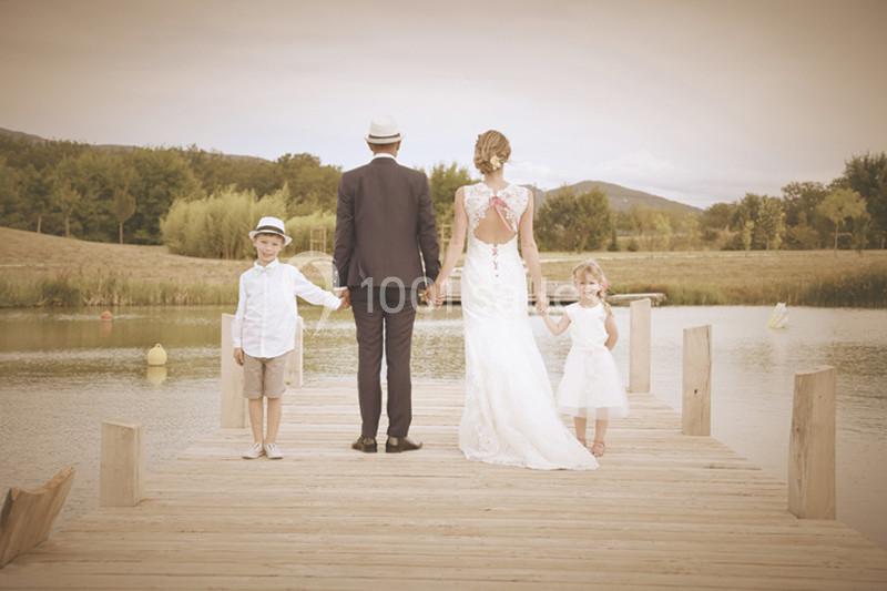 Une famille en tenue élégante, debout sur un ponton en bois, face à un lac et un paysage naturel.