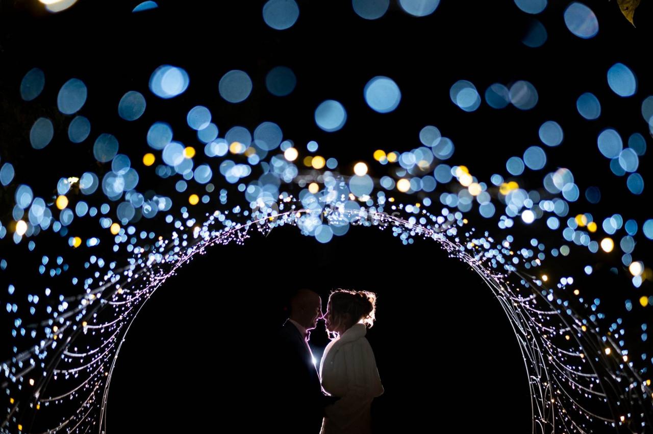 Deux silhouettes se tenant face à face sous une arche illuminée de lumières floues dans un décor nocturne.