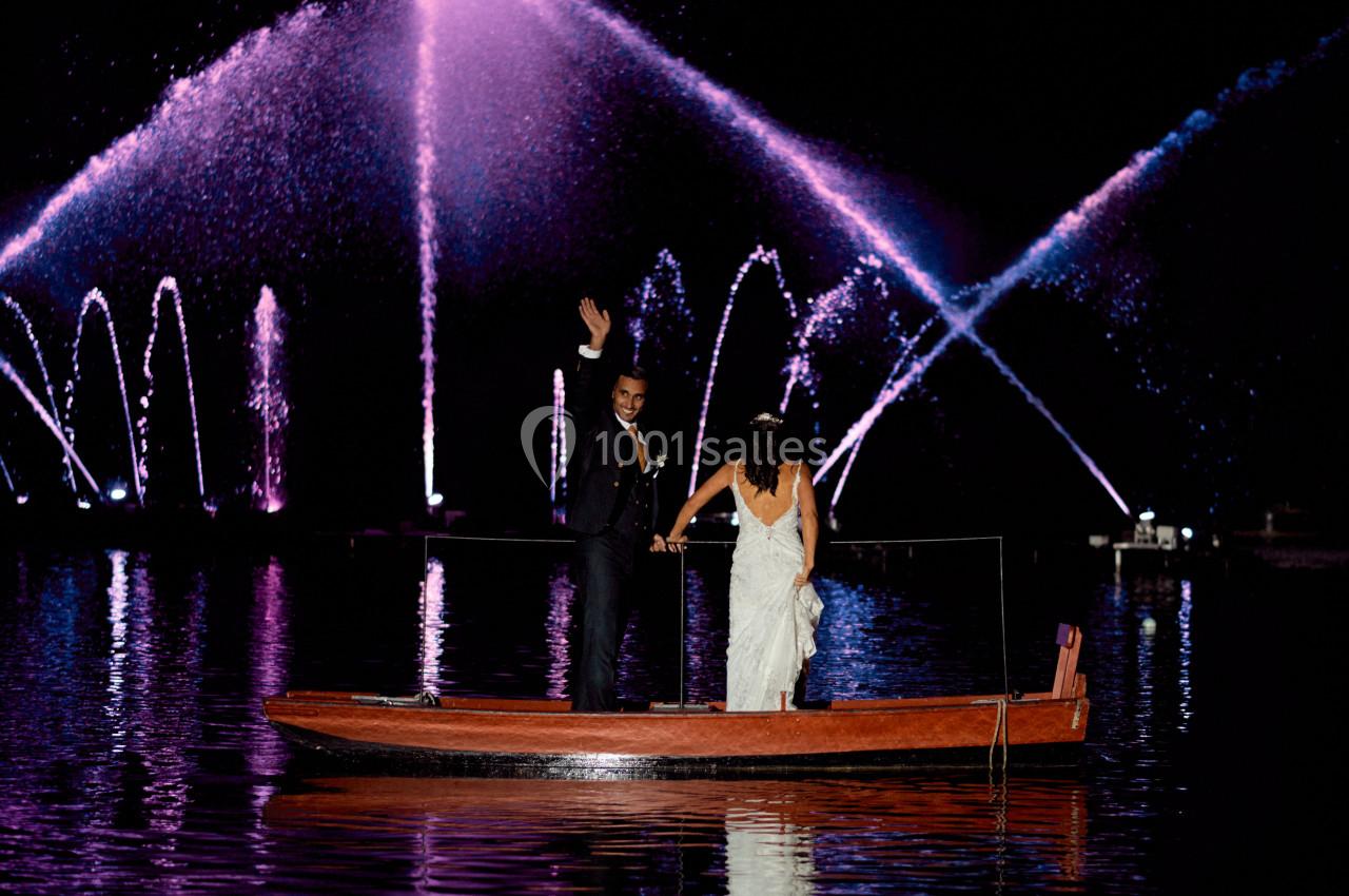 Un couple en tenue de mariage sur une barque, avec des fontaines lumineuses violettes en arrière-plan, de nuit.
