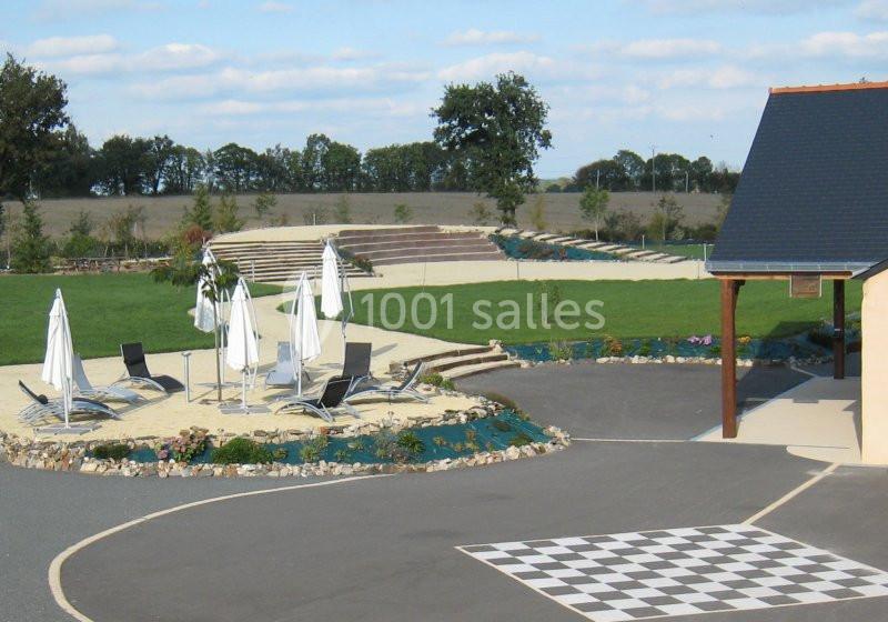 Espace extérieur avec chaises longues et parasols sur du sable, entouré de pelouse et d'un bâtiment à droite.