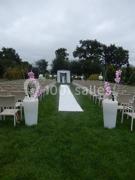 Allée centrale bordée de chaises et décorée de fleurs menant à une arche de mariage en extérieur sur pelouse.