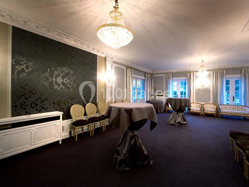 Salle élégante avec lustres, tables hautes drapées, chaises classiques et grandes fenêtres laissant entrer la lumière…