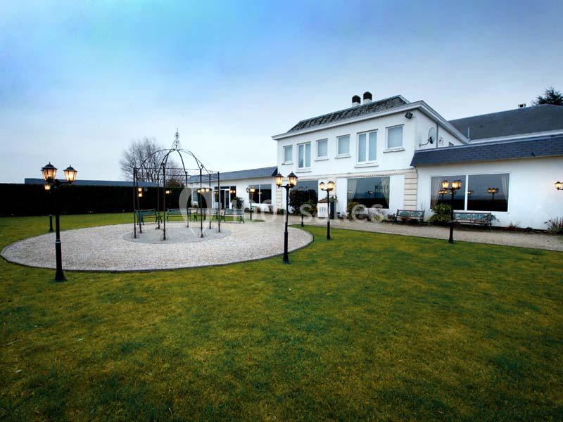 Grande maison blanche avec jardin, pelouse, allée circulaire en gravier et lampadaires allumés au crépuscule.