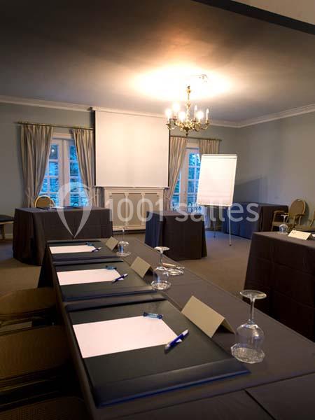 Salle de réunion avec tables disposées en U, blocs-notes, stylos, verres et un tableau de présentation devant des rideaux.