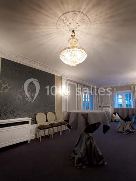 Salle élégante avec murs ornés, lustre en cristal, tables hautes drapées de tissu gris et chaises alignées.